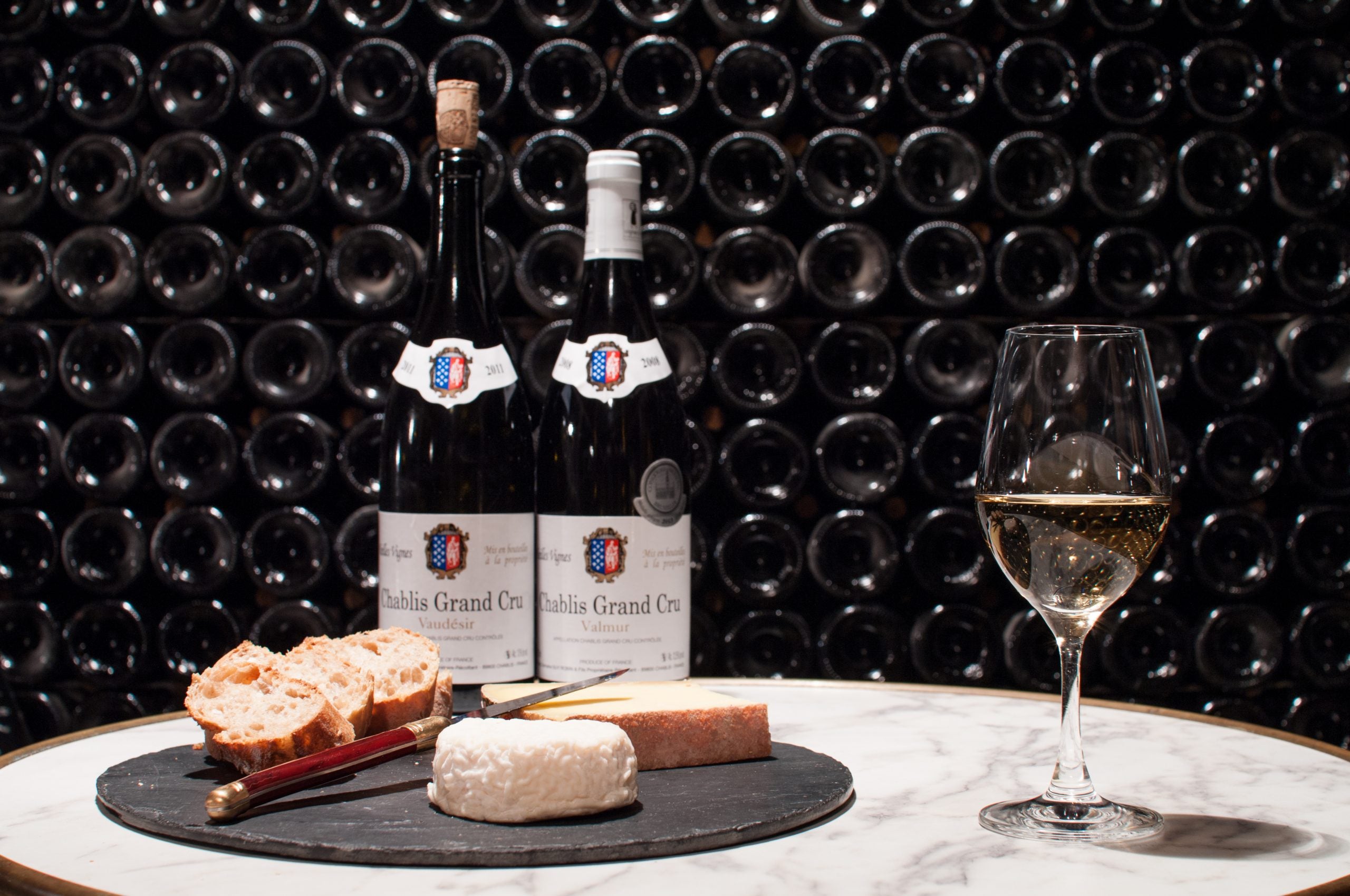 Two bottles of wine with a glass and cheese platter on a table against a wall of wine bottles.