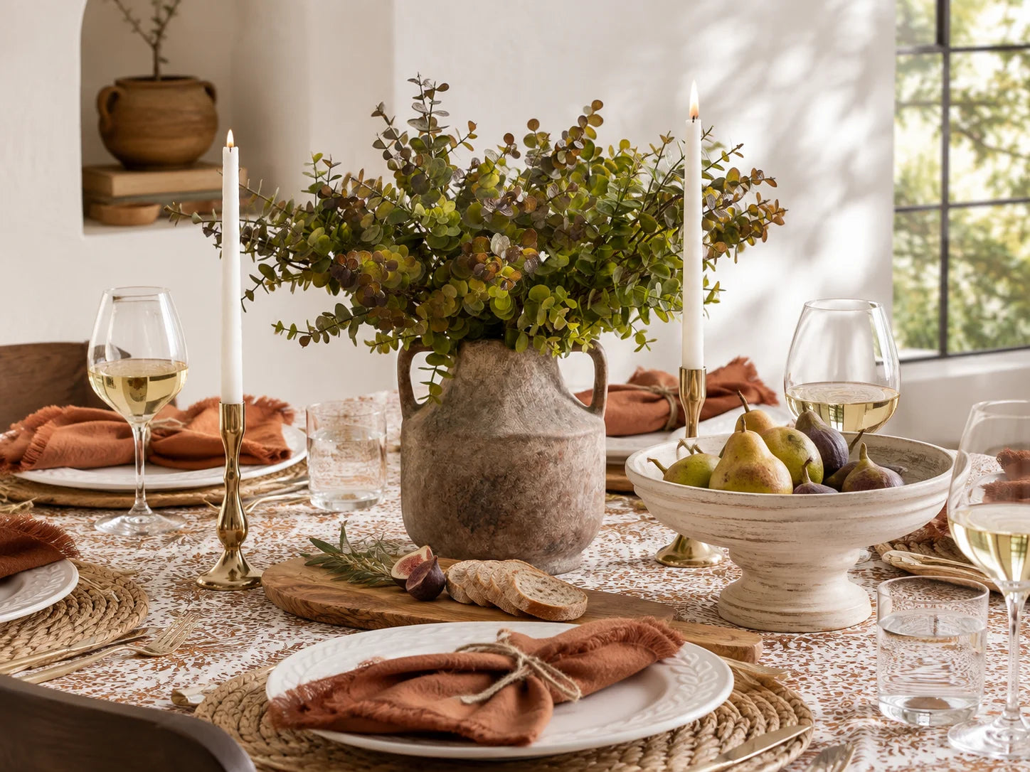 A styled dining table centerpiece featuring a rustic ceramic vase filled with green foliage, flanked by tall white taper candles in brass holders, with plates, wine glasses, bread, and a bowl of pears in a warm, sunlit room.