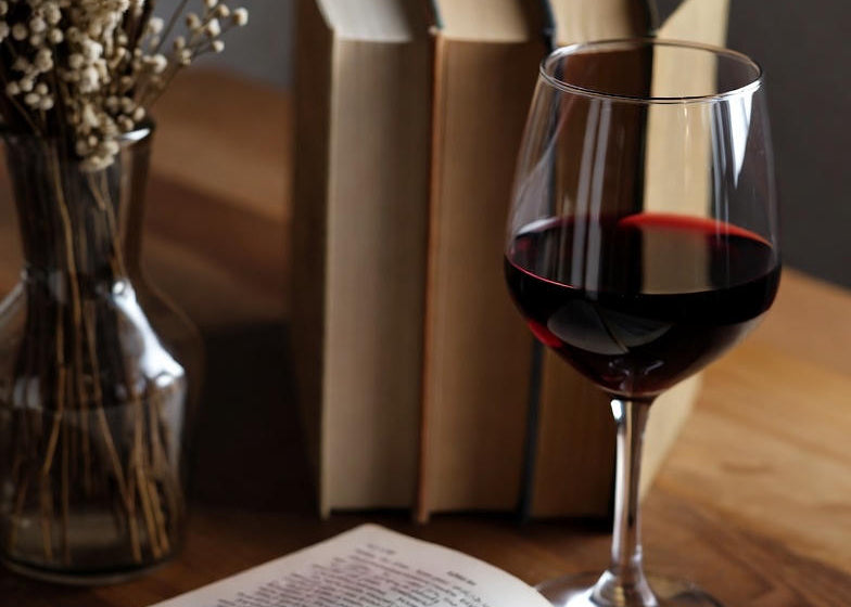 Glass of red wine next to an open book on a wooden surface with books and a vase in the background.